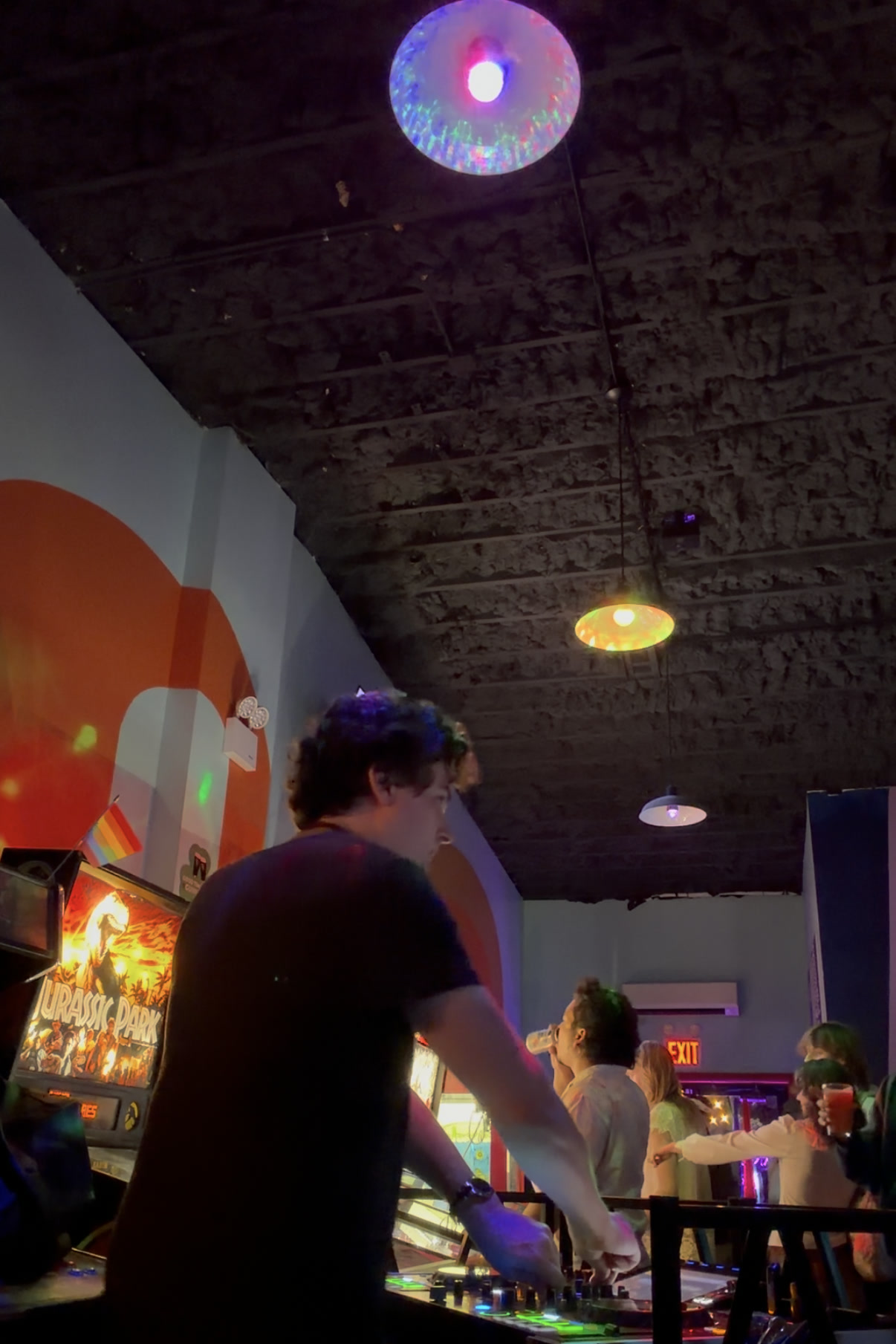 Looking out across the decks to the crowd, drinks in hand, arcade cabinets and a Jurassic Park pinball machine lit up behind them