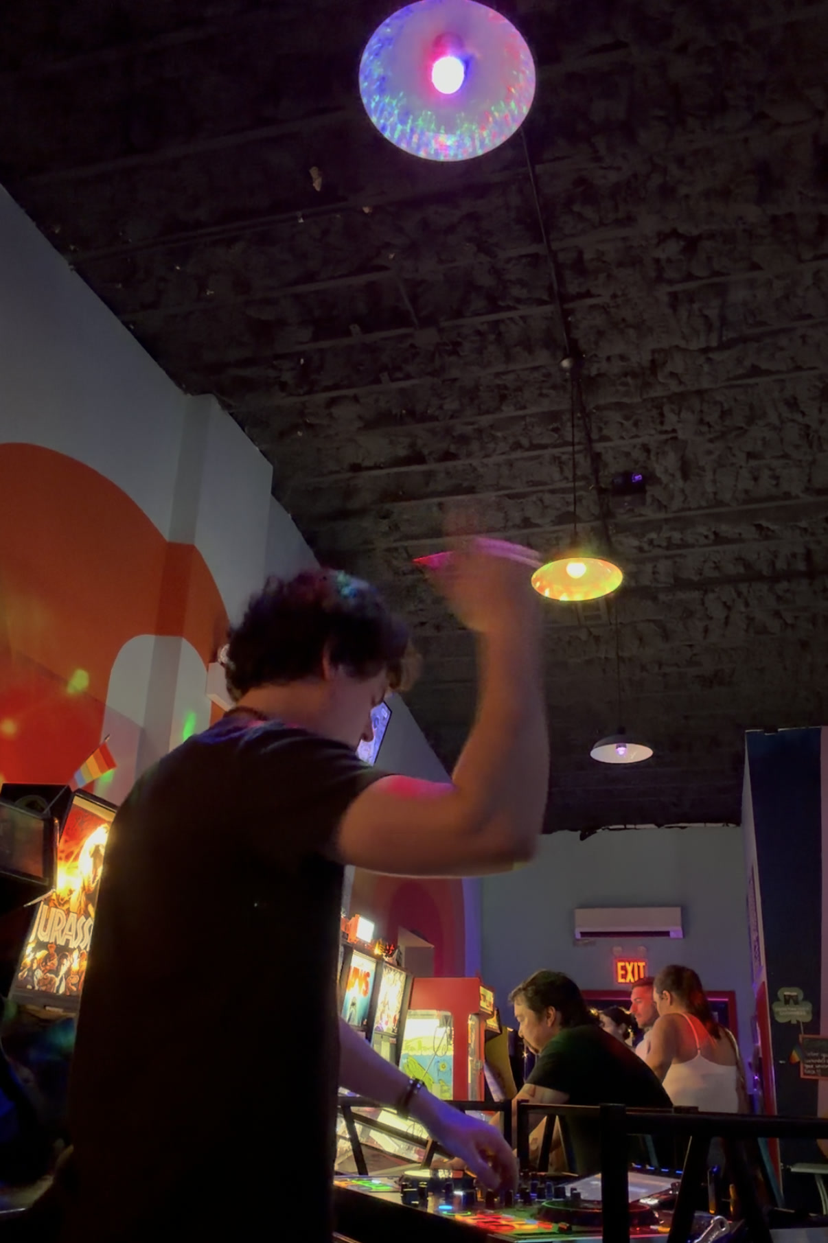 Playing the decks at the Happy Valley Arcade Bar under a mirrorball, one hand up, pinball machines lined along the wall