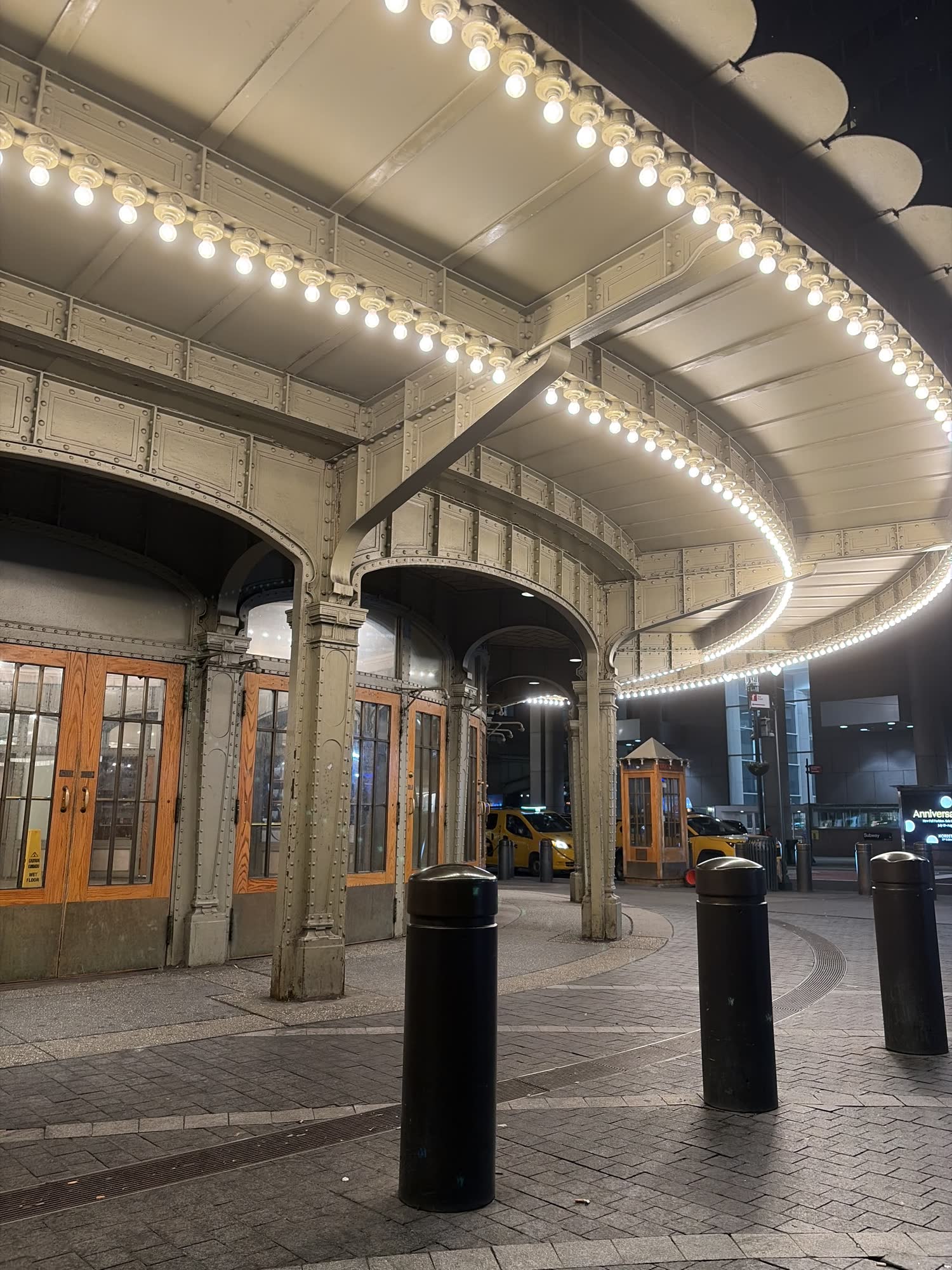 A riveted iron canopy strung with bare bulbs over the station entrance at night, cabs at the curb