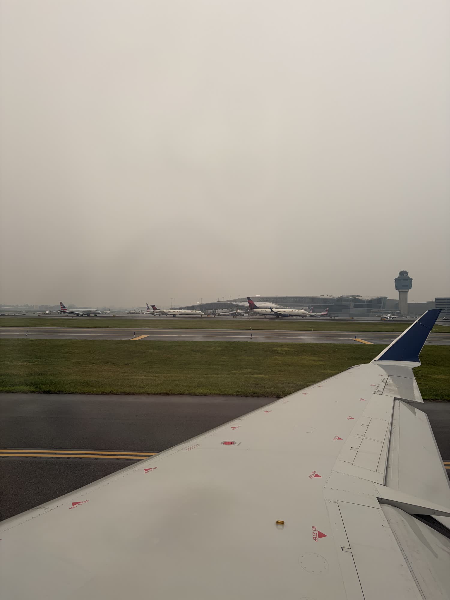 Over the wing on the ground at LaGuardia, storm-grey sky, the control tower and a line of planes waiting in the distance