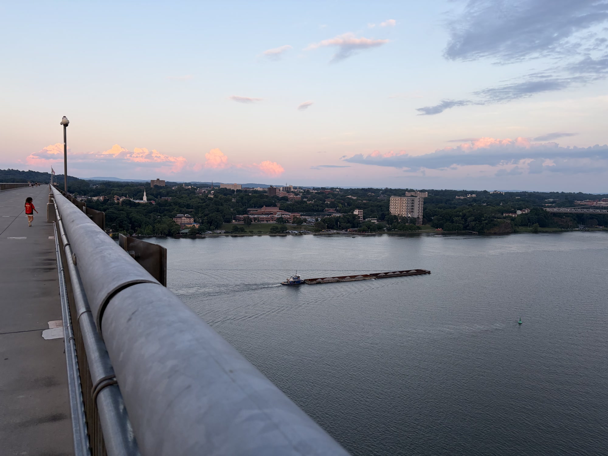 A barge and tug on the Hudson at sunset, seen from a bridge walkway, pink clouds over the far bank