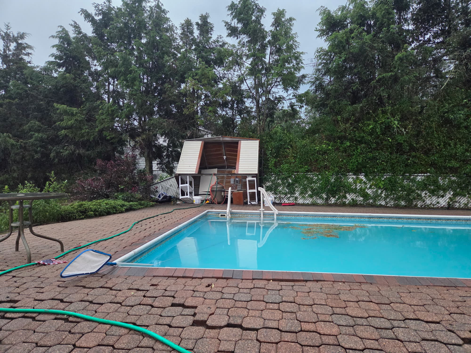 Pool shed lifted off its footing and tilted over, siding peeled back and windows sitting on the ground, beside a clear blue pool in the rain